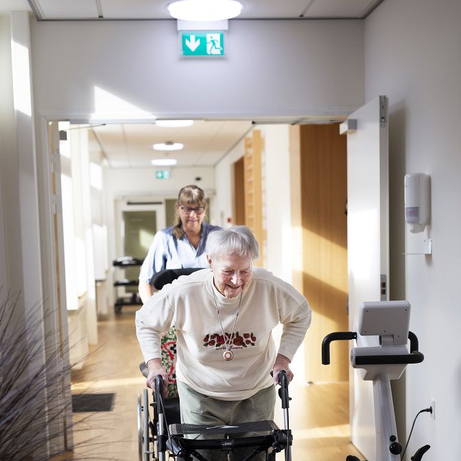 Ældre dame går ned ad en gang. Der er en SOSU-assistent, som følger efter og hjælper hende.