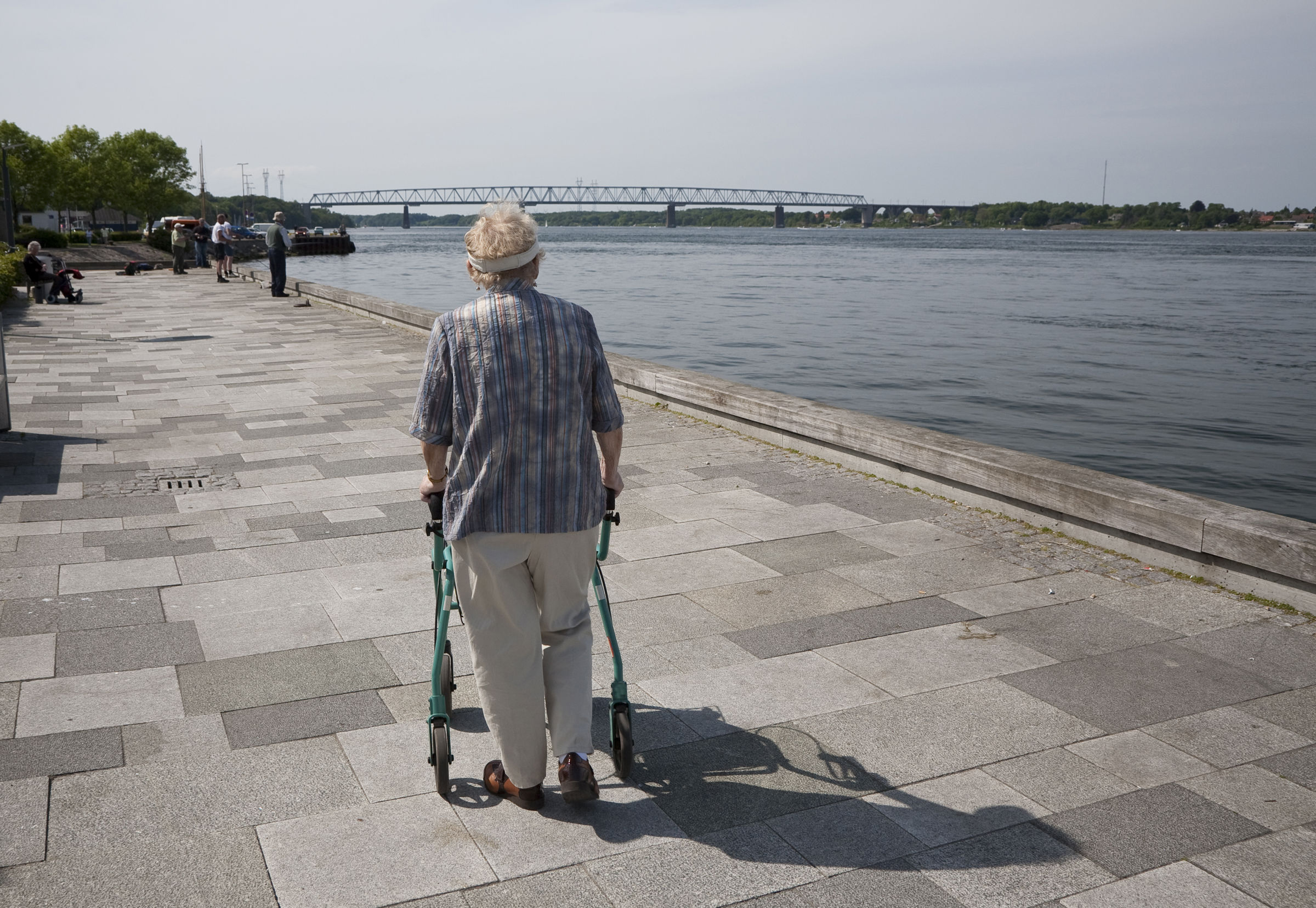 Ældre person med rollator går en tur ved vandet. Man kan se en bro i distancen.